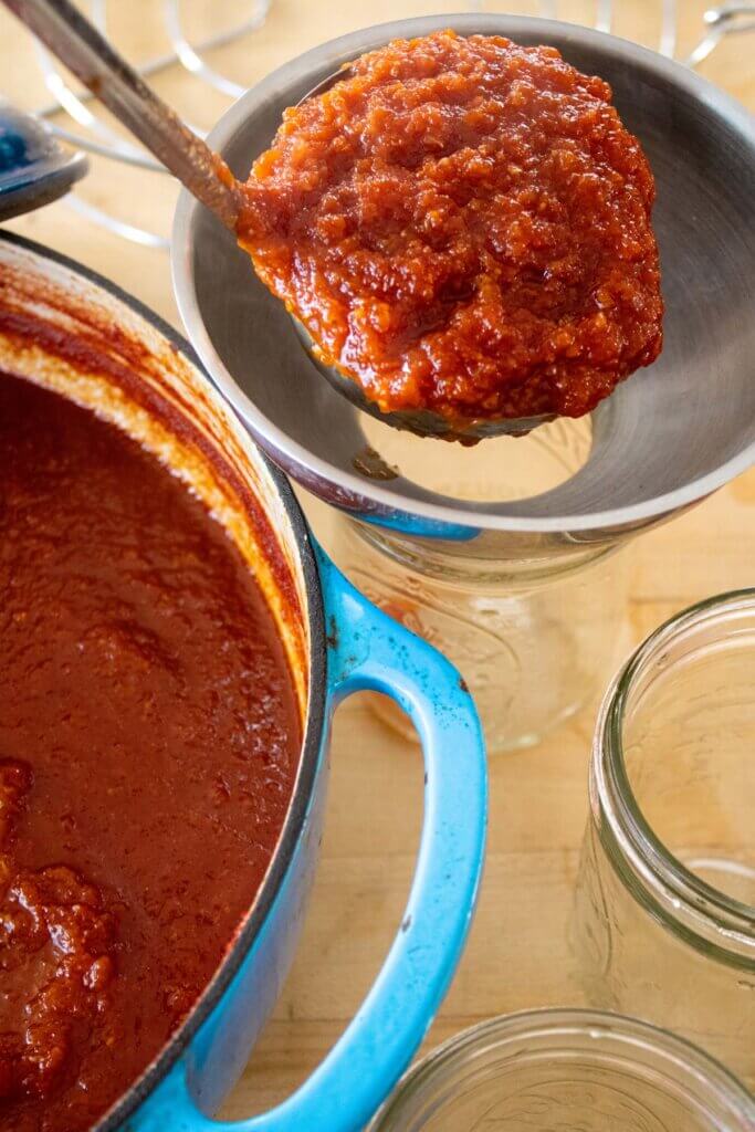 A ladle of BBQ sauce filling a canning jar prepped with a funnel.
