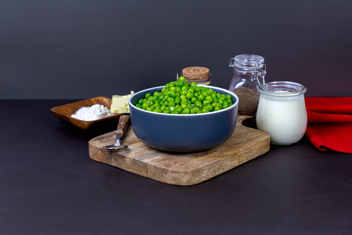 A bowl of fresh peas surrounded by creamed peas ingredients: flour, butter, and cream.