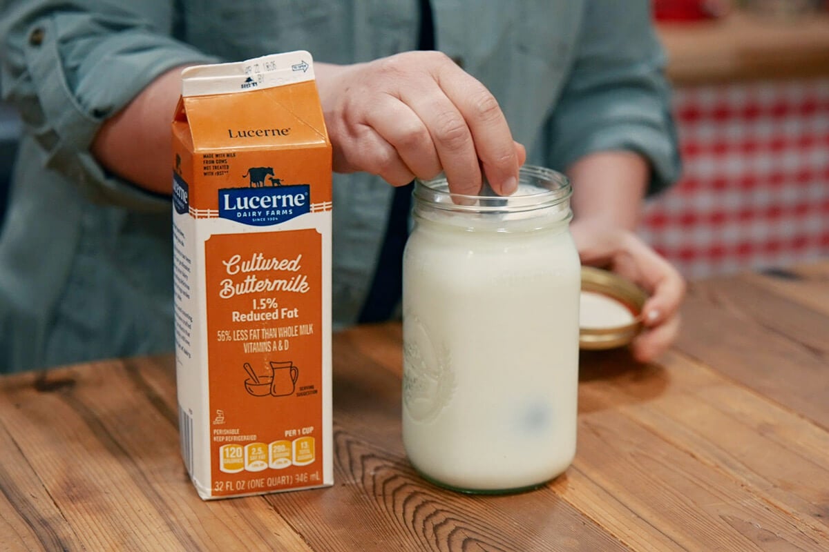 Hand holding a spoon stirring buttermilk and milk together in a jar.