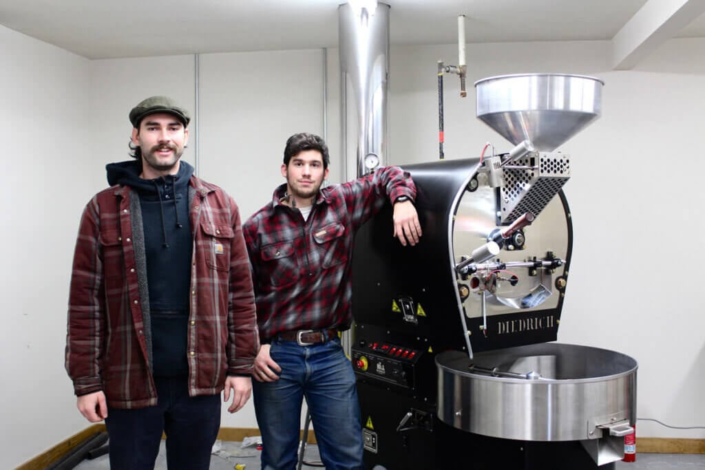 Two men standing beside a coffee roaster.