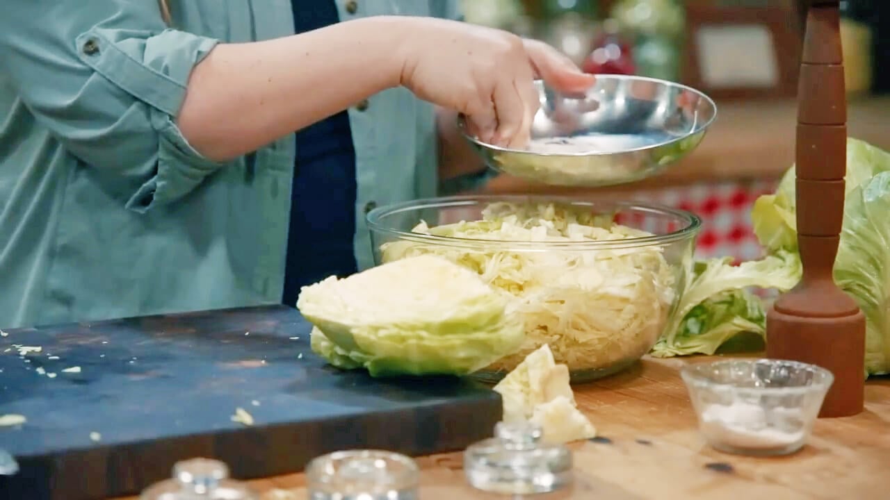 Hands sprinkling salt over shredded cabbage in a bowl.