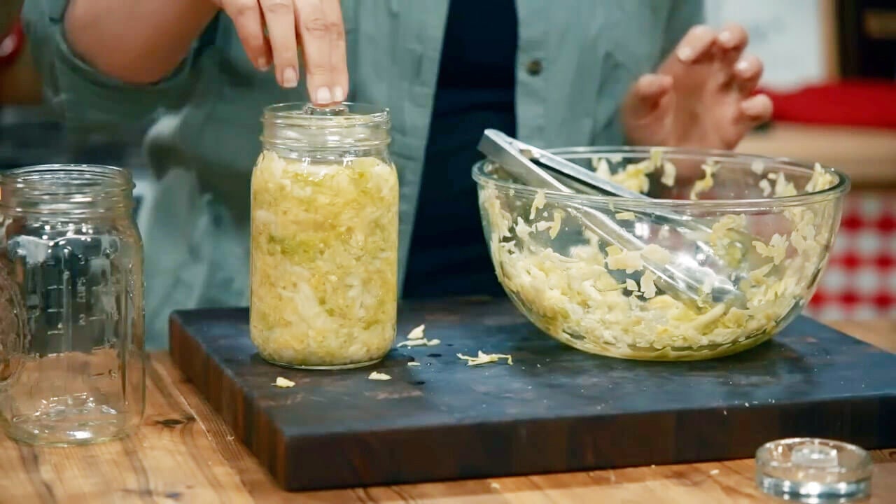 A hand placing a fermenting weight on top of jar filled with shredded cabbage.