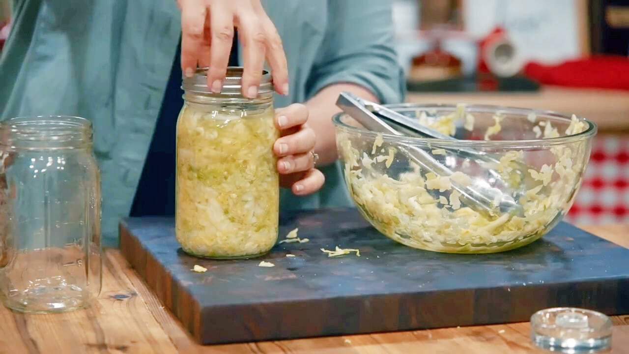 Hand placing a lid on a glass jar filled with shredded cabbage.