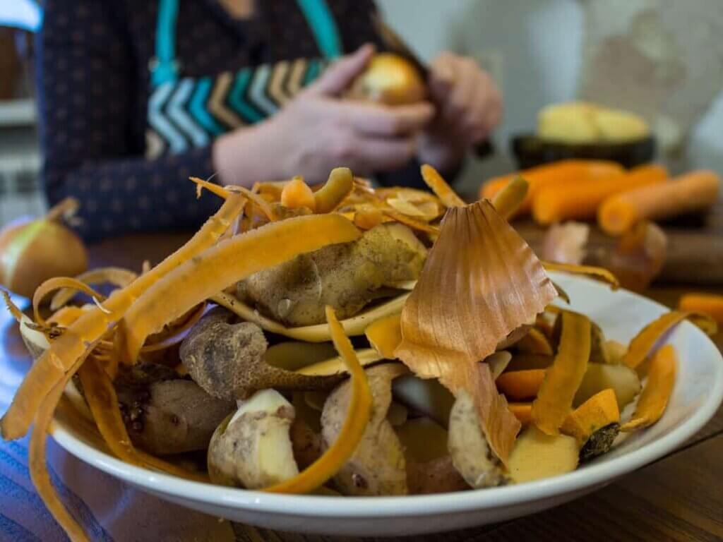A plate of vegetable scraps on a table with a woman in the background peeling vegetables.