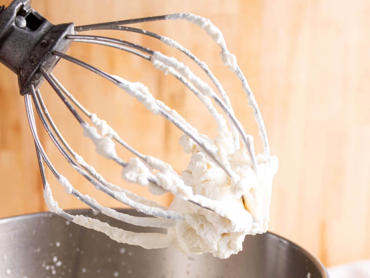 A stand mixer with the whisk attachment lifted to show the texture of whipped cream.