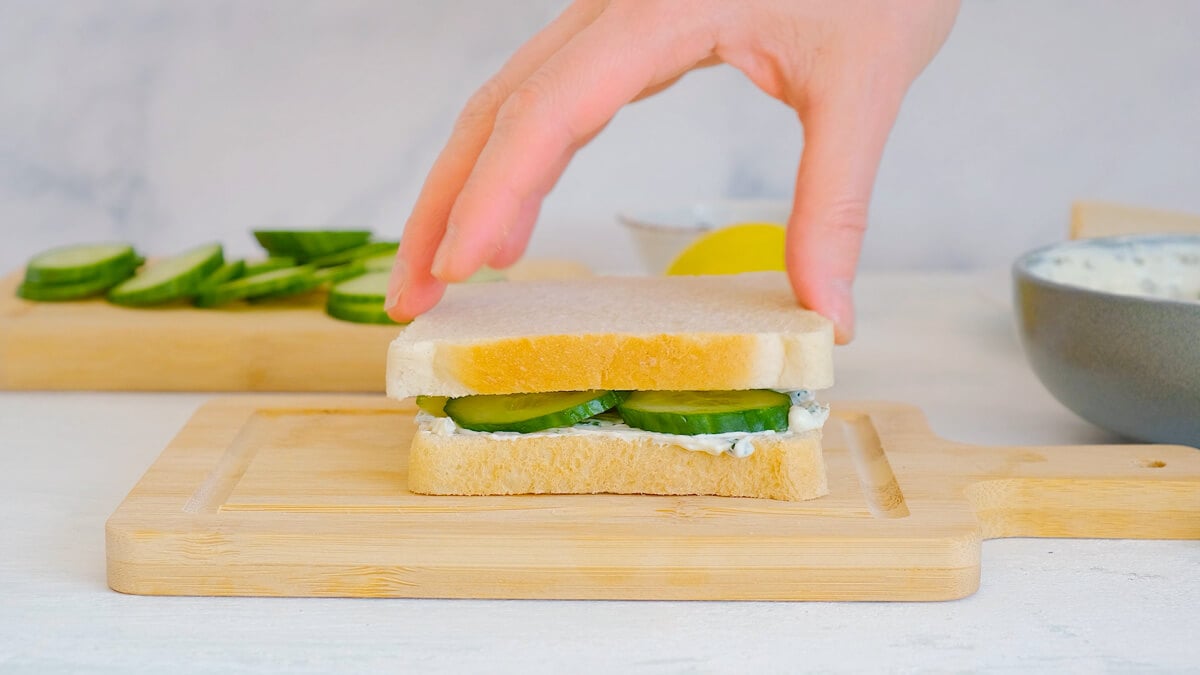 A hand placing a top slice of bread onto a cucumber sandwich.
