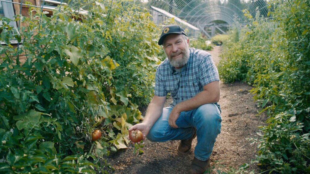 A man crouched in the garden holding a tomato.
