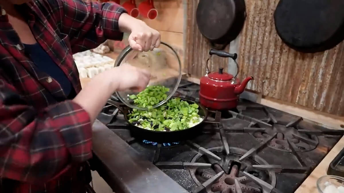 Hands pouring chopped from a bowl into a cast iron skillet filled with saut&eacute;d onion.