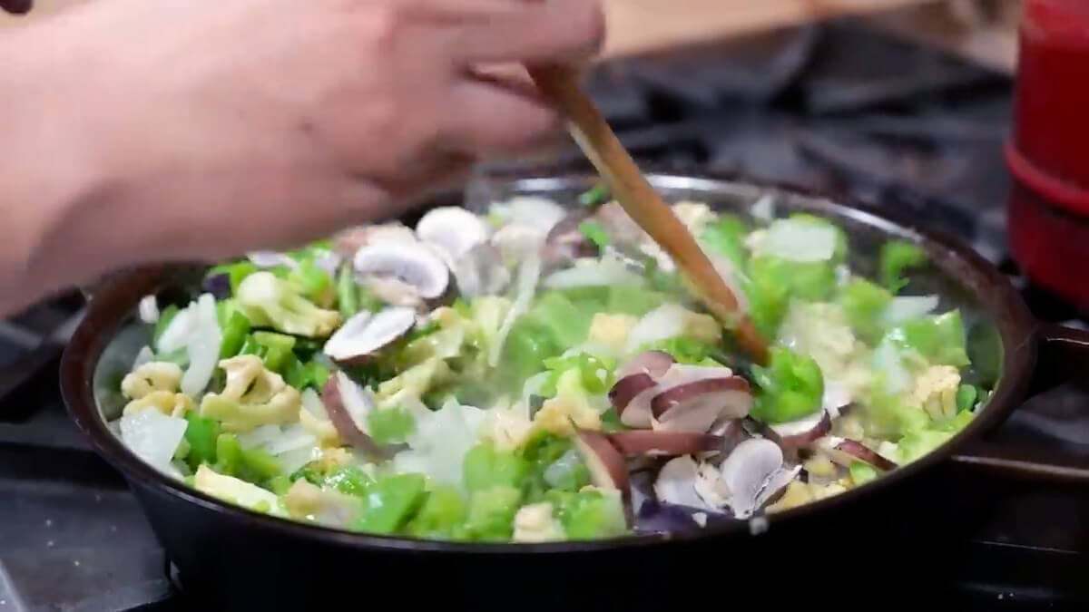 A hand holding a wooden spoon stirring saut&eacute;d vegetables in a cast iron skillet.
