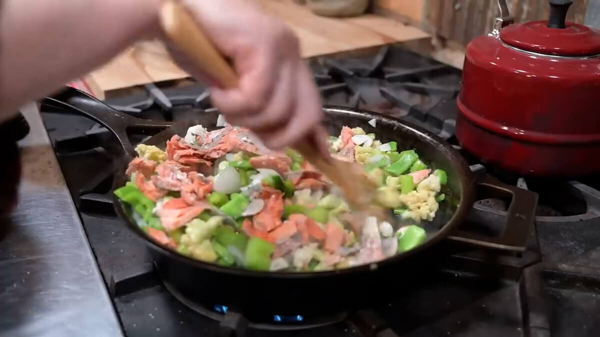 A hand holding a wooden spoon stirring saut&eacute;d vegetables and cooked salmon in a cast iron skillet.