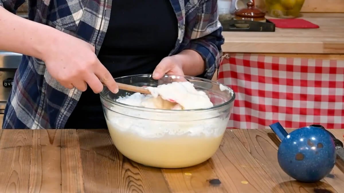 Hands using a spatula to gently fold egg whites into eggnog in a bowl.