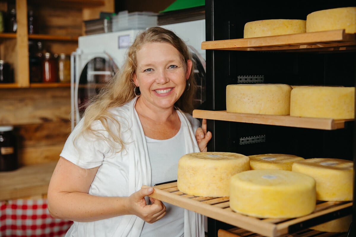A woman next to a cheese cave with multiple wheels of homemade cheese inside.