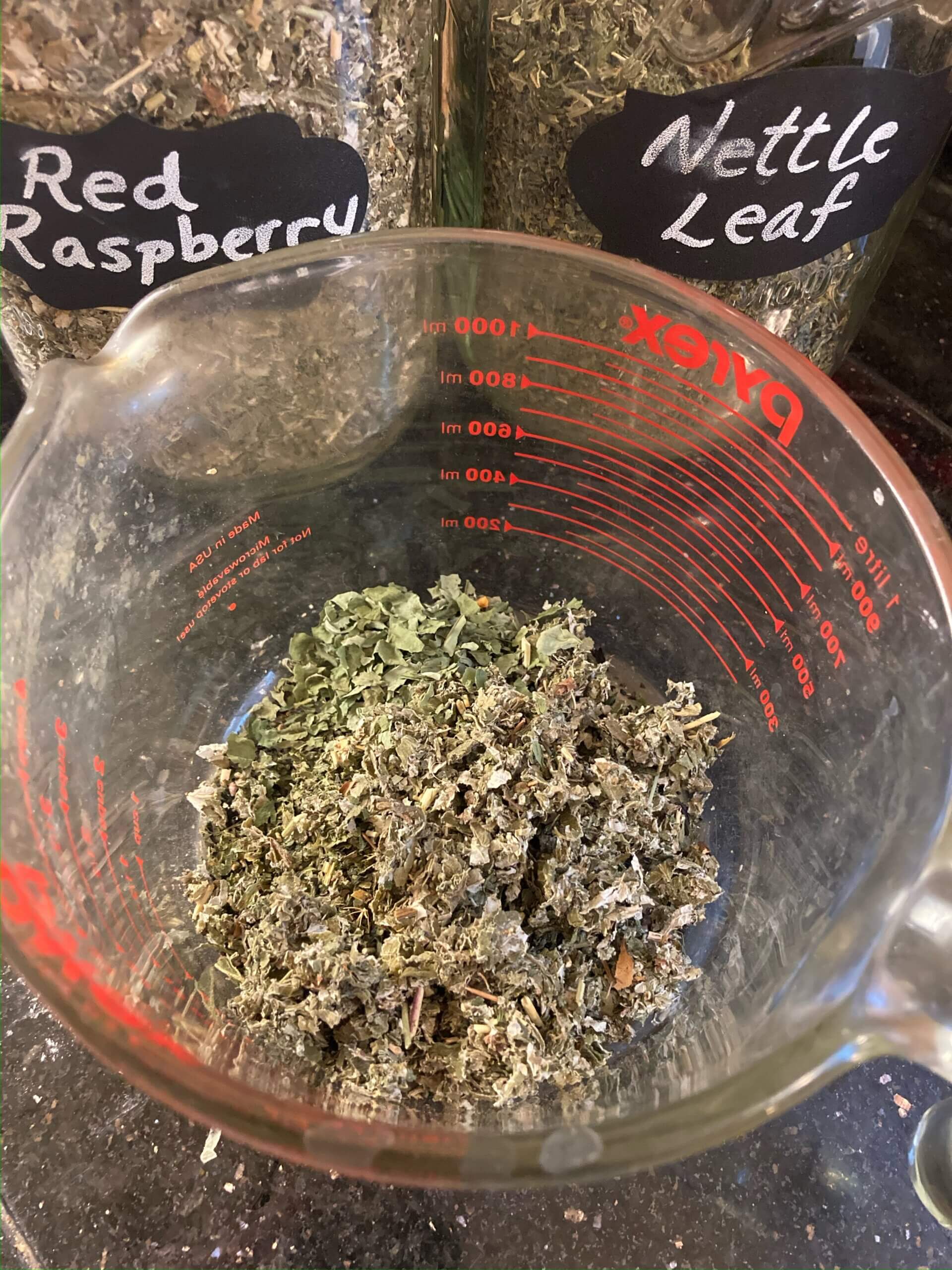 Dried herbs in a measuring cup with jars of herbs in the background.