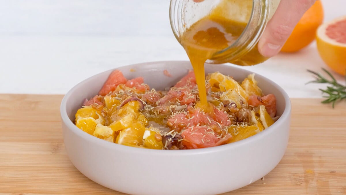 Hands pouring citrus dressing from a jar onto citrus salad in a white bowl.