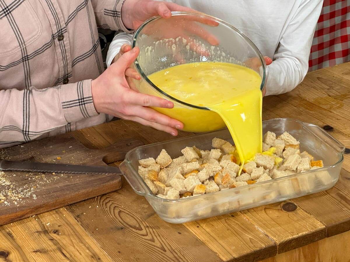 A bowl of custard being poured over a pan of bread cubes.