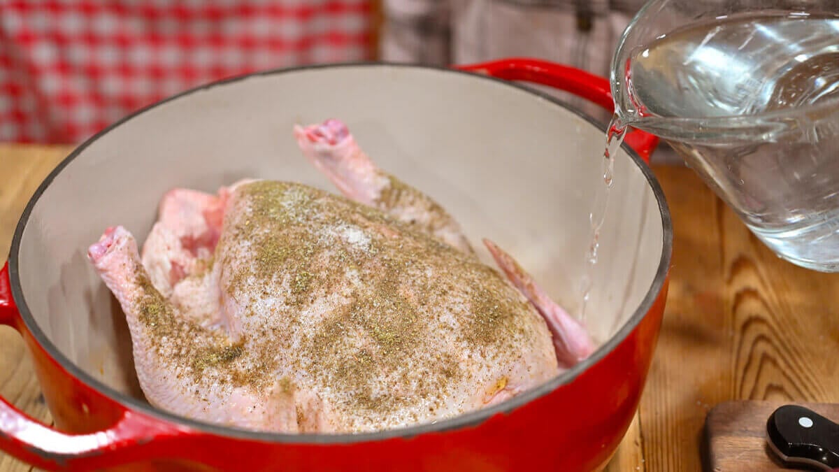 A whole chicken in a Dutch oven with water being added to the bottom of the pot.