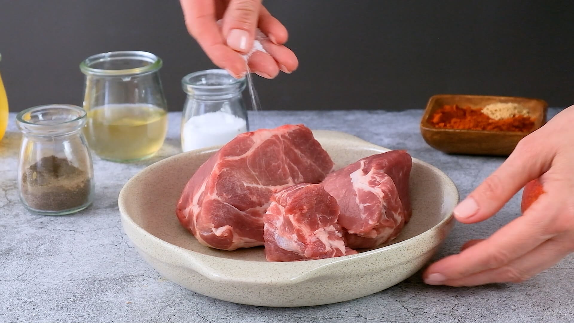 Hands sprinkling salt over a raw pork shoulder.