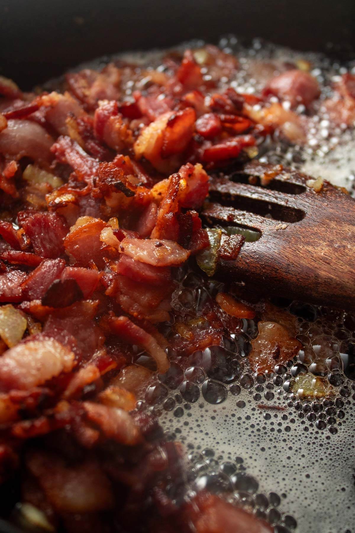 A spatula stirring bacon and onions that are frying in a pan.