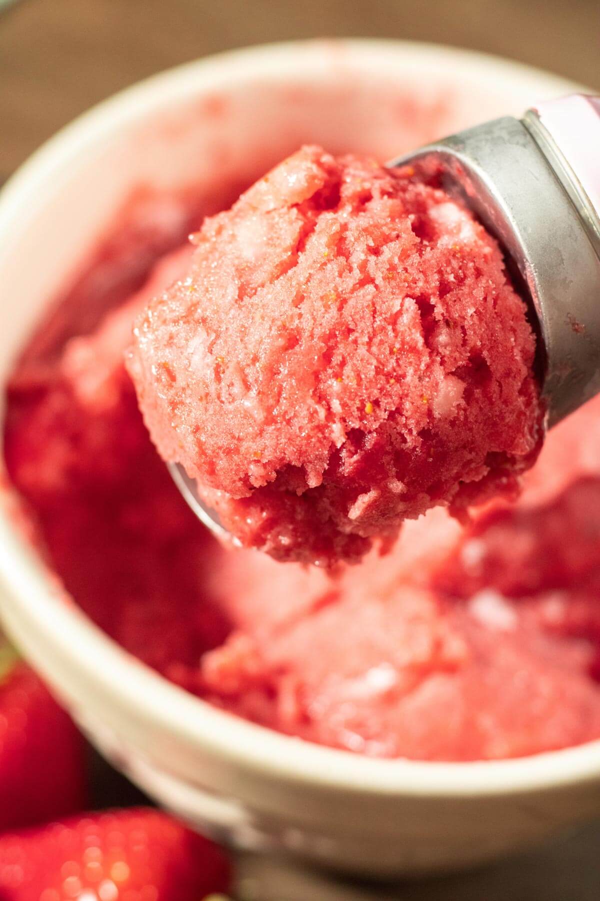 A close up shot of a white bowl filled with strawberry sorbet, and a scoop removing a serving to show texture.