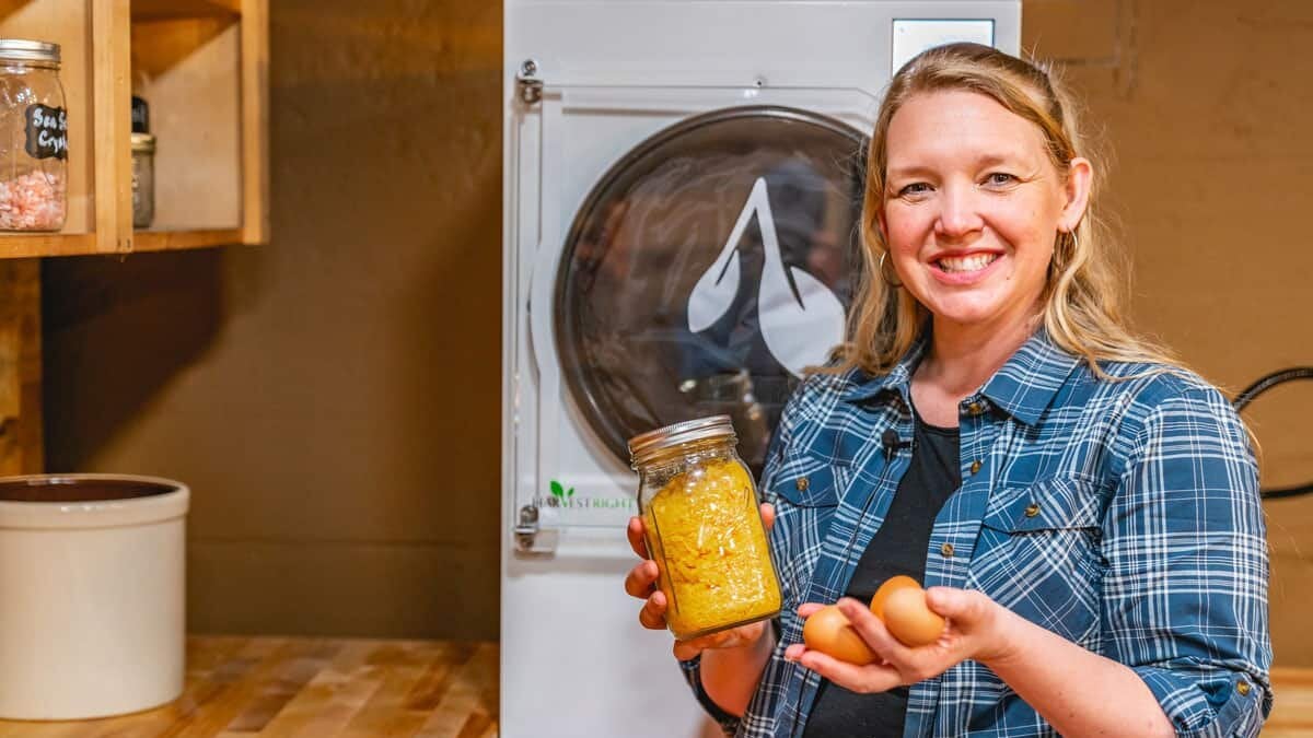 A woman standing in front of a harvest right freeze dryer holding a jar of powdered eggs in one hand and farm fresh eggs in the other hand.