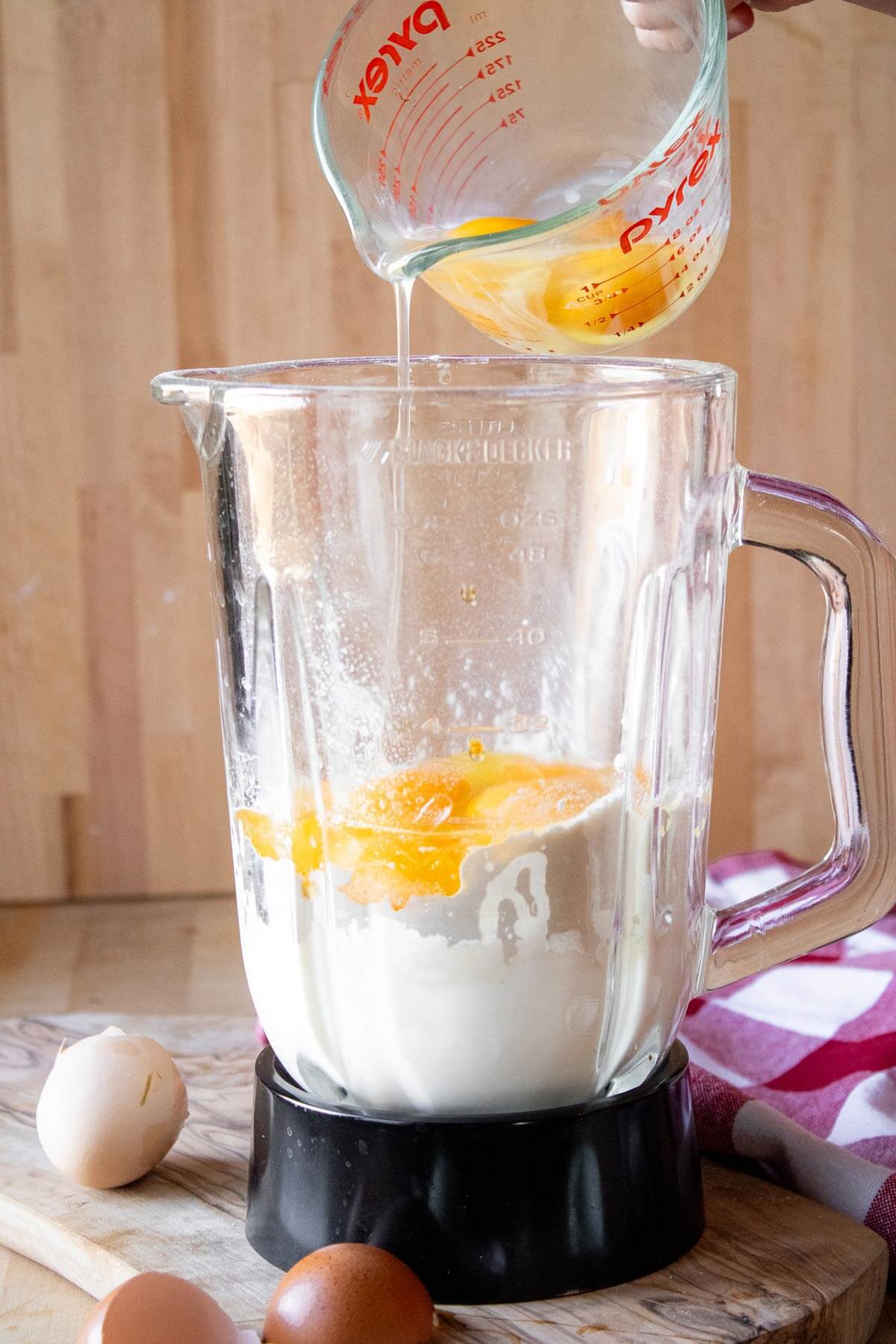 A measuring cup pouring eggs into a blender filled with milk and flour.