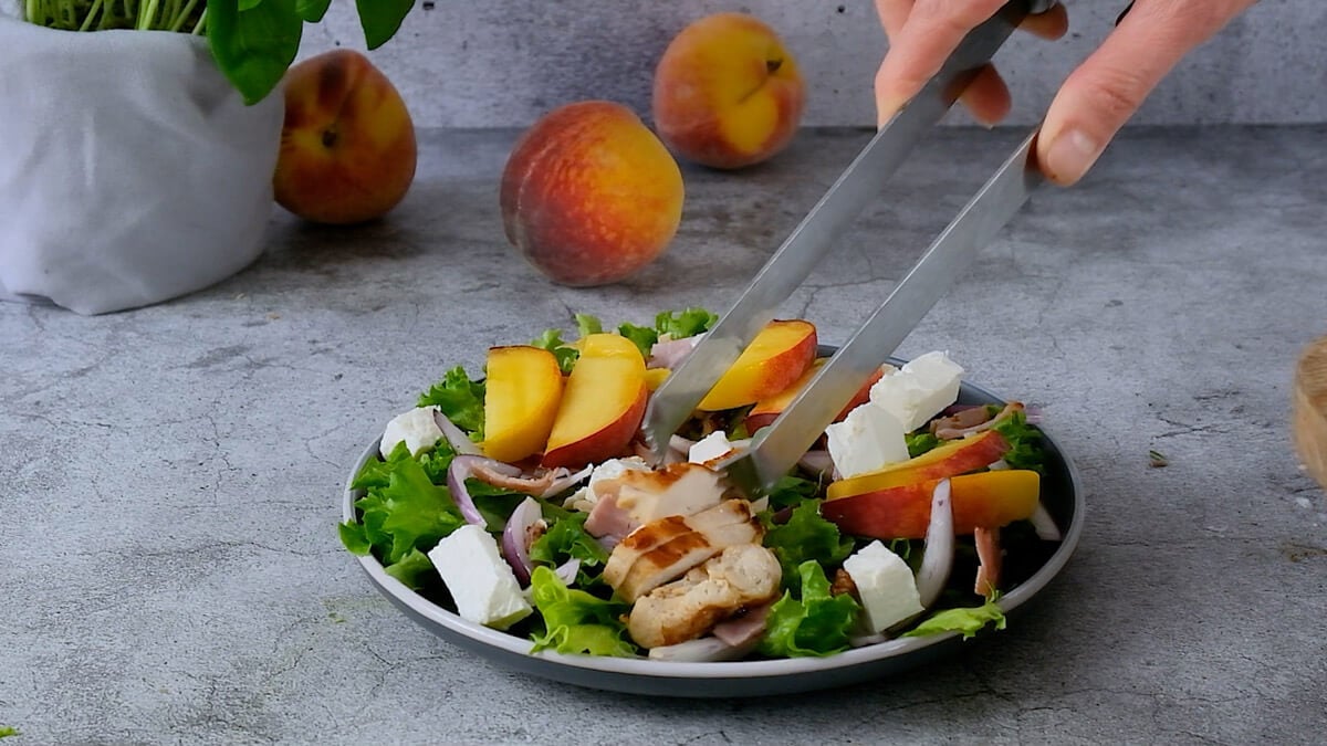 Tongs arranging peach salad with feta on a plate.