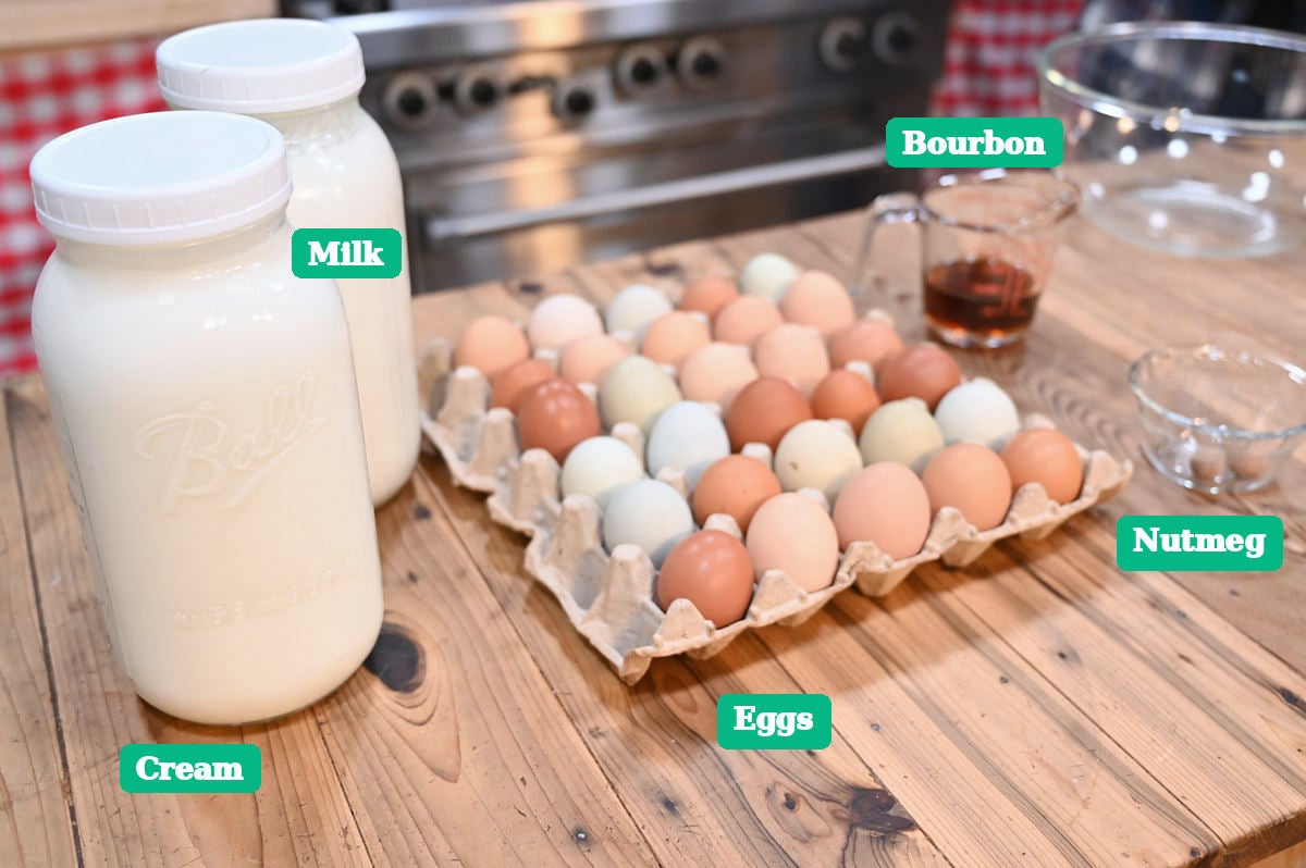 Cream, milk, eggs, bourbon, and nutmeg arranged on a wooden counter for aged eggnog.