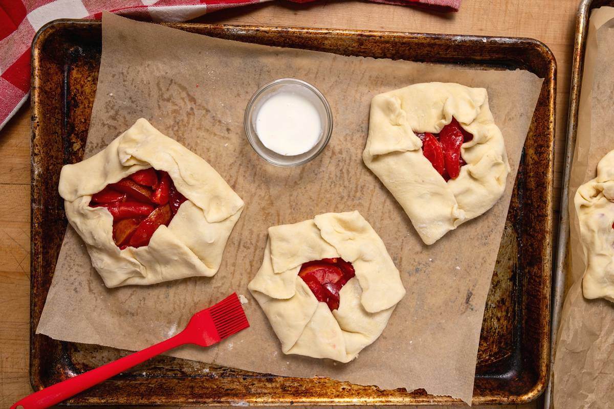 Unbaked plum pies on parchment with cream and a red pastry brush.