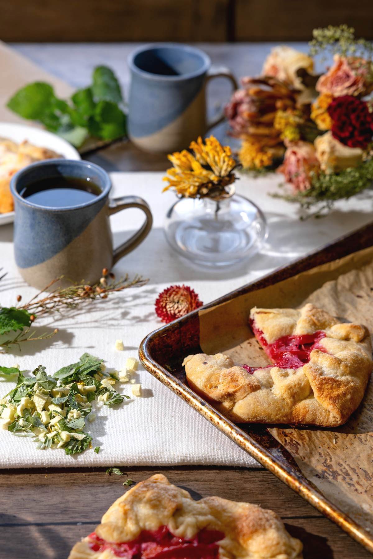 Plum pies on a baking sheet beside coffee mugs and autumn flowers.