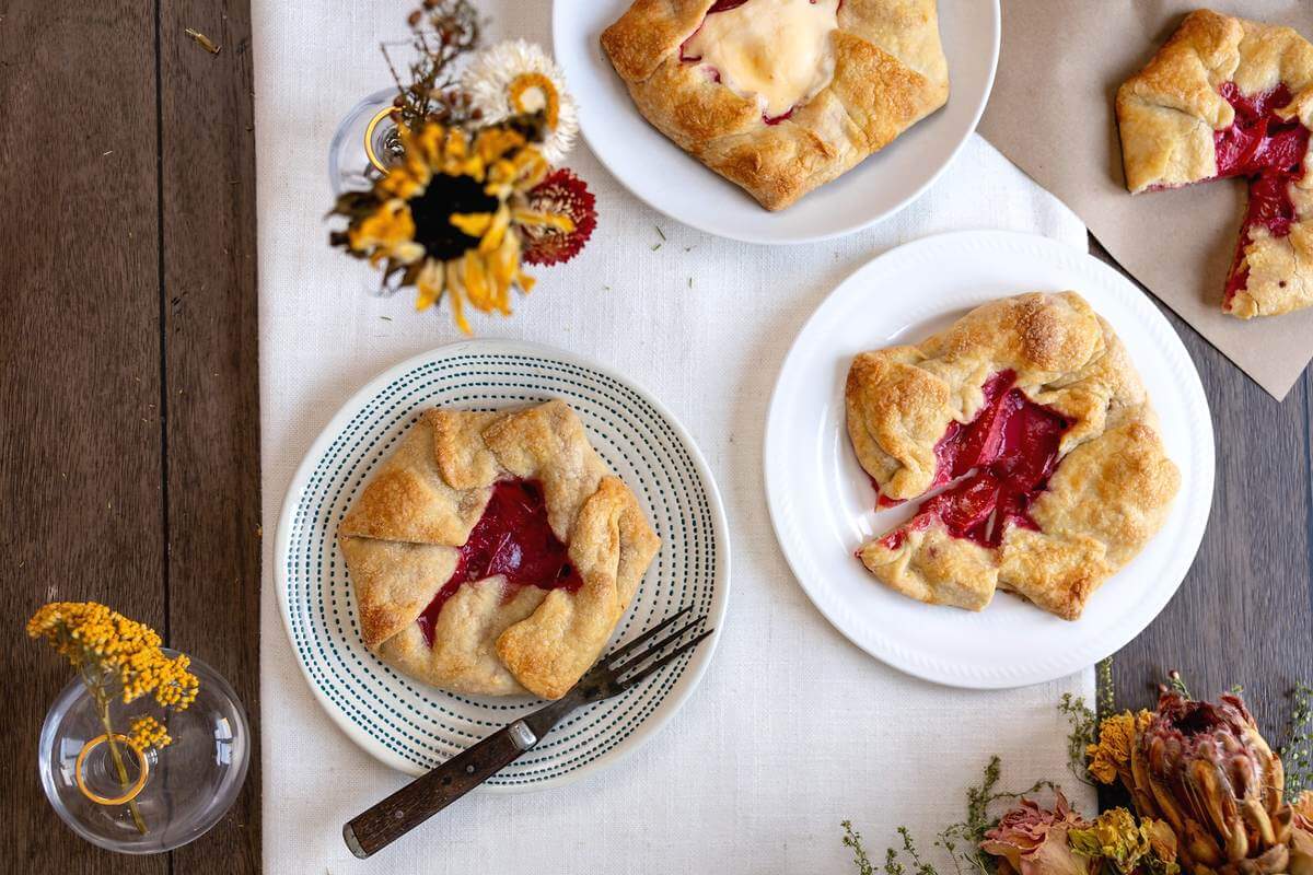Two plum hand held pies on patterned and white plates beside flowers.