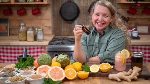A woman holding a dropper bottle of herbal bitters and fresh produce and herbs in front of her.