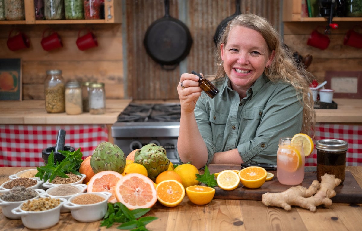 A woman holding a dropper bottle of herbal bitters and fresh produce and herbs in front of her.