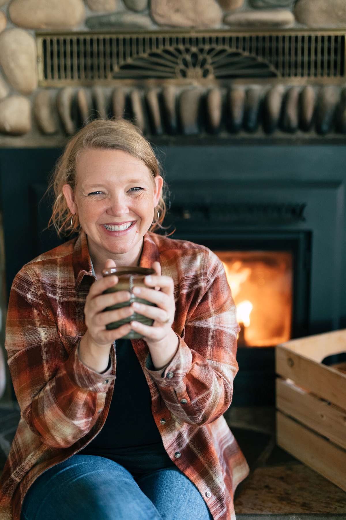 Woman in a plaid shirt holding a ceramic mug beside a glowing fireplace