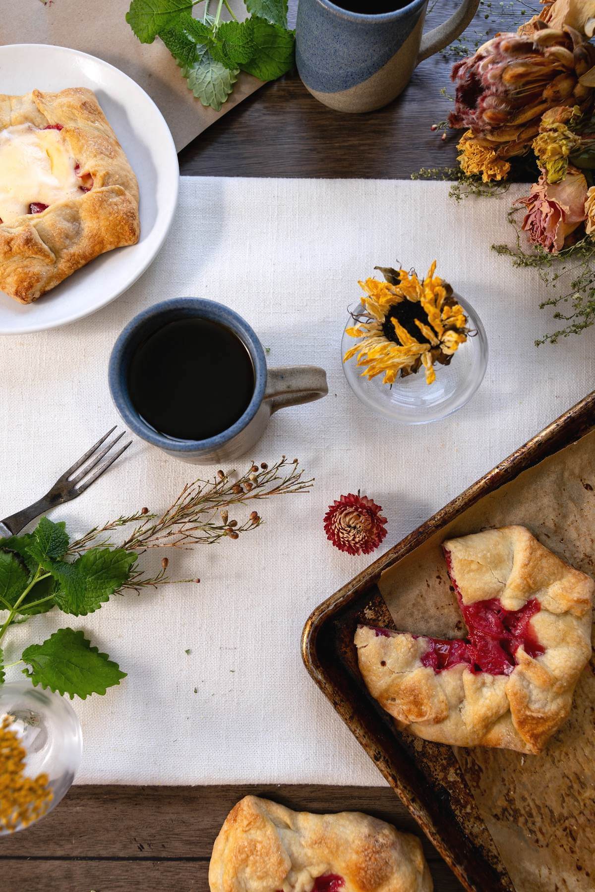 Plum pies on a plate and baking sheet beside coffee and autumn flowers.