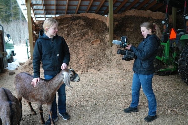 A young girl with a camera filming another young girl with a goat.