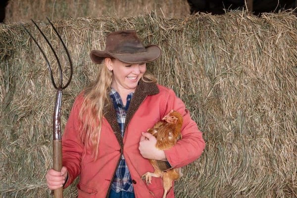 A woman holding a pitch fork in one hand and a chicken in the other with a stack of hay in the background.