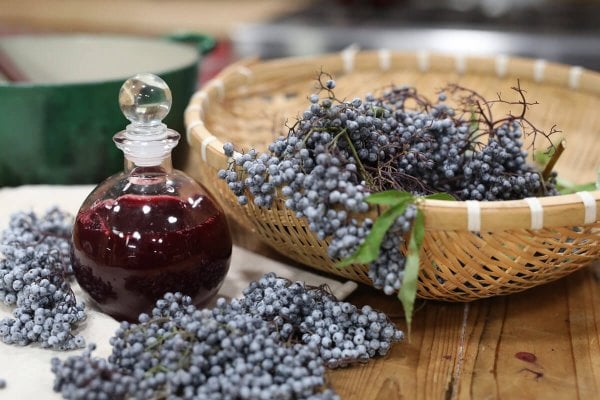 A basket full of fresh elderberries sitting next to a bottle of homemade elderberry syrup.