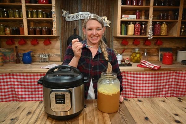 A woman holding a spoon of gelled broth with a gallon of broth and an instant pot on the counter in front of her.