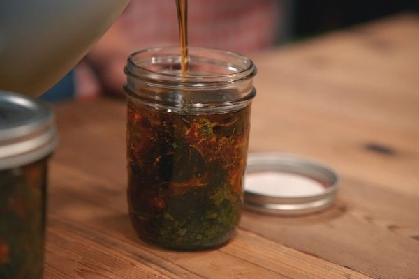 Pouring oil over dried herbs in a glass jar.
