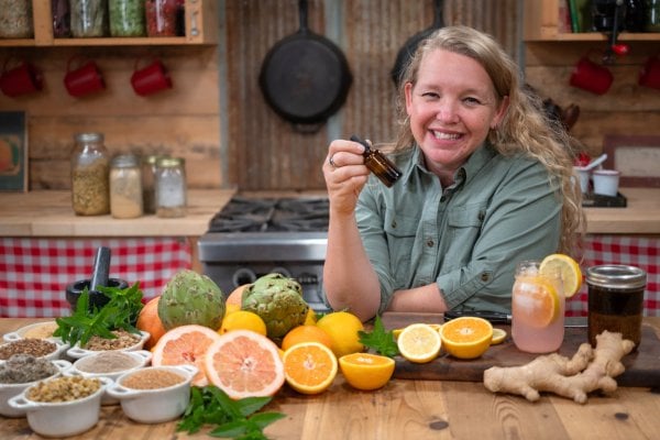 A woman holding a dropper bottle of herbal bitters and fresh produce and herbs in front of her.