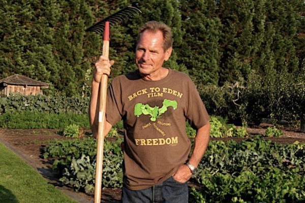 A man standing in front of a garden leaning on a rake.